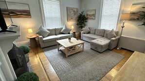 A modern living room with two sofas, a coffee table, and a floor lamp. There are two windows with blinds, a painting on the wall, and a small plant on the coffee table. - Spacious Suite 1 Bed/ 1 Bath/ Pullouts Sofas - Unit 5 1 Bedroom Apts (Newport)