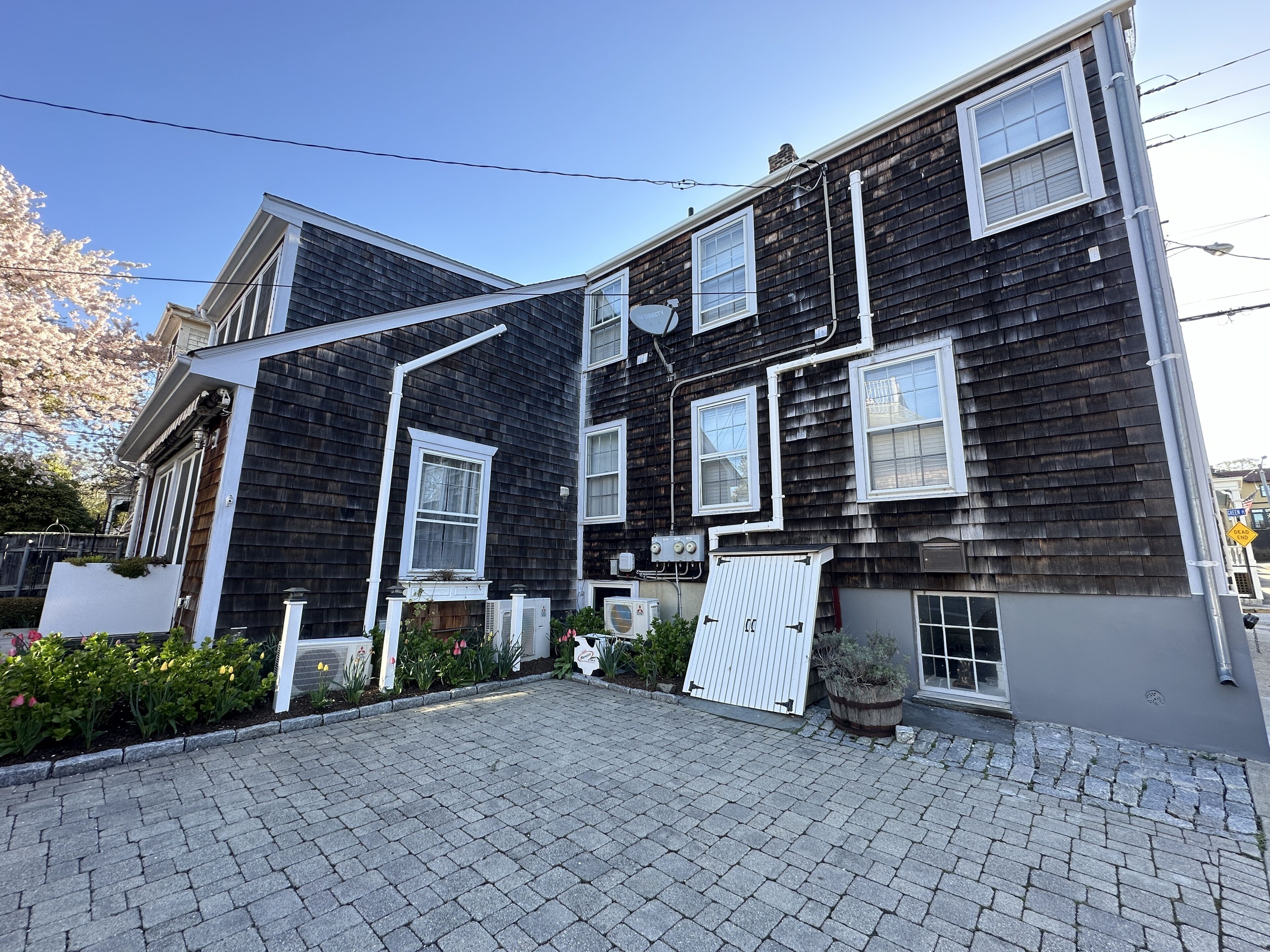 A two-story building with a dark exterior and white trim. There are multiple windows, some with shutters, and a garage door. The ground is paved with cobblestones.