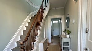 Een hal met een houten trap, een radiator, een vloermat, een console tafel met een plant, en een deur.