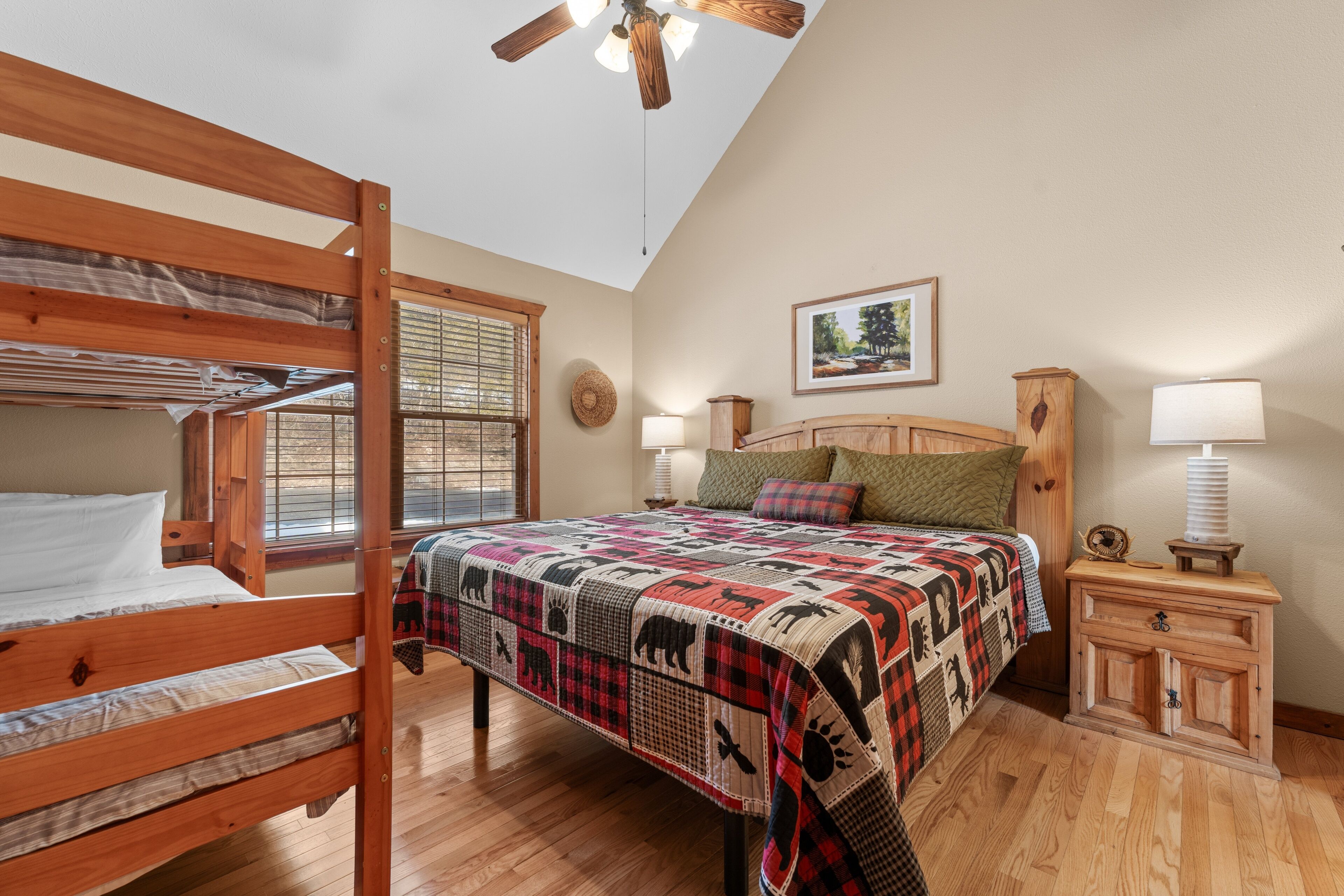 Une chambre avec un lit superposé, un grand lit recouvert d’une couette à motifs, une tête de lit en bois, une table de chevet avec une lampe et une fenêtre avec des stores.