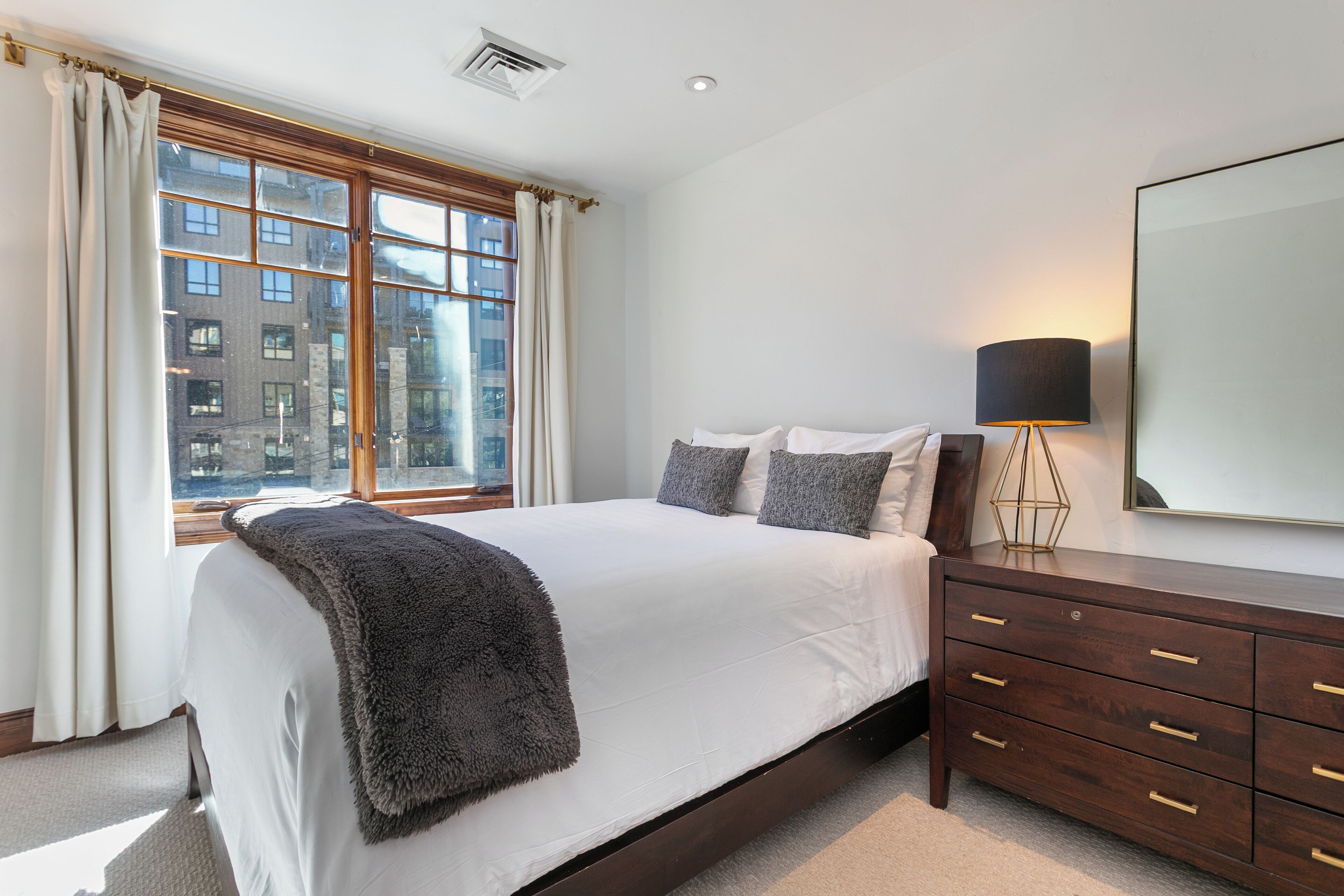 A bedroom with a large bed, a dresser with drawers, a lamp, and a window with curtains.