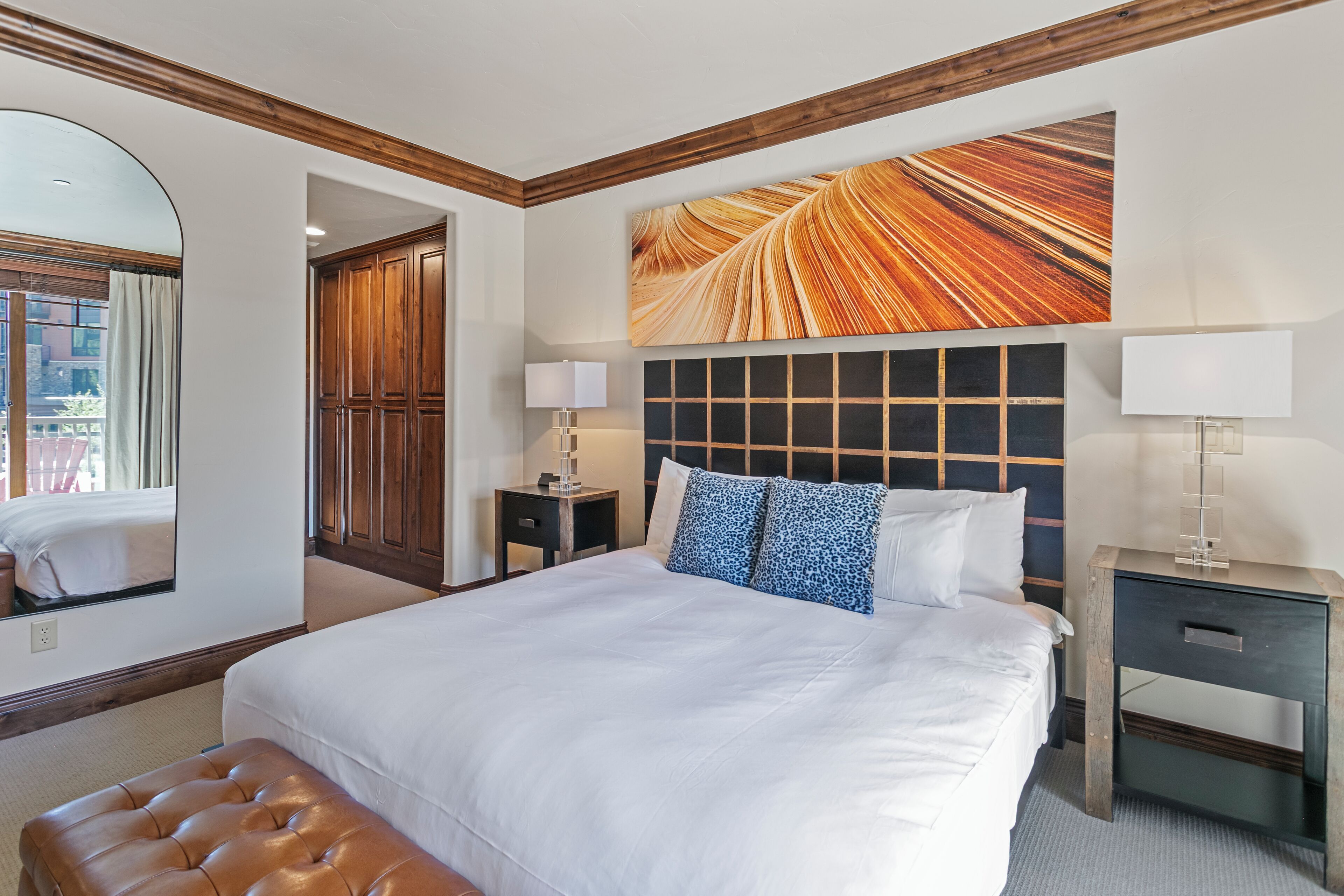 A hotel room with a large bed, a wooden headboard, a mirror, a bedside table with a lamp, and a view of the outside through a window.