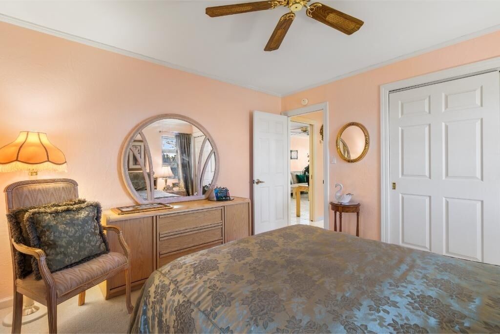 A bedroom with a bed, a dresser, a chair, a ceiling fan, and a door leading to another room.