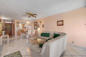 A living room with a sofa, coffee table, and chairs. There is a dining area with a table and chairs. The room has a ceiling fan and a chandelier. - Menehune Shores, #301 2 Bedroom Condo (Kihei)