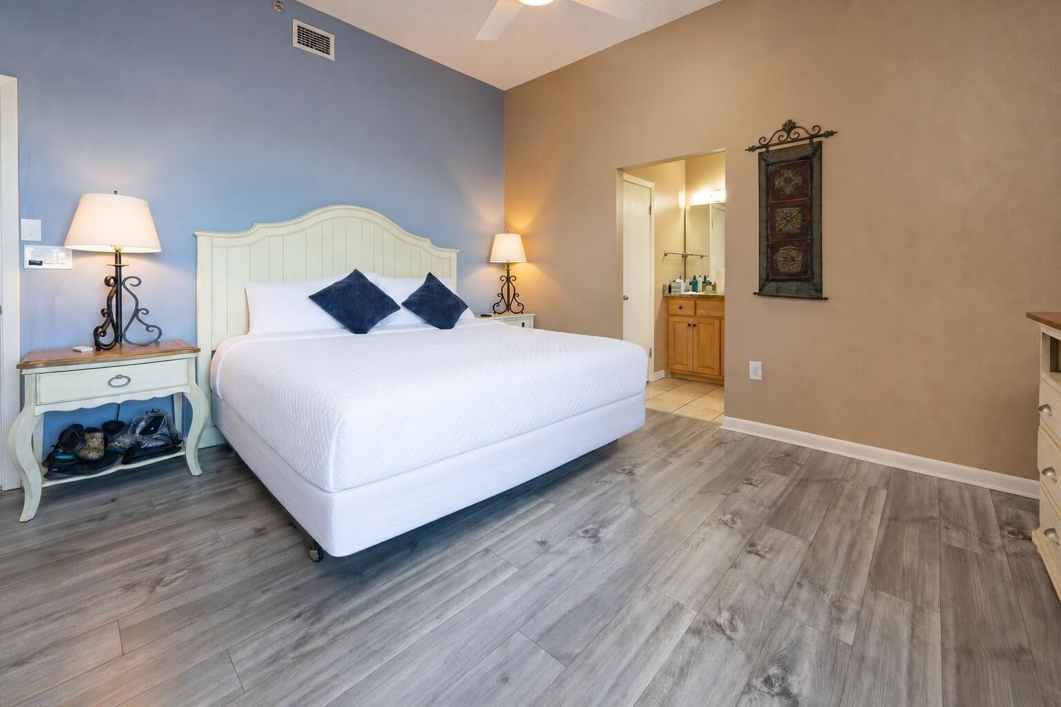 A bedroom with a bed, two bedside tables with lamps, a wooden floor, a door leading to a bathroom, and a mirror on the wall.