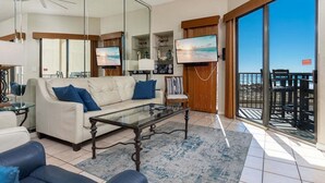 Ein geräumiges Wohnzimmer mit einem weißen Sofa, einem Glastisch und einem großen Fernseher. Ein Balkon bietet Blick aufs Meer.