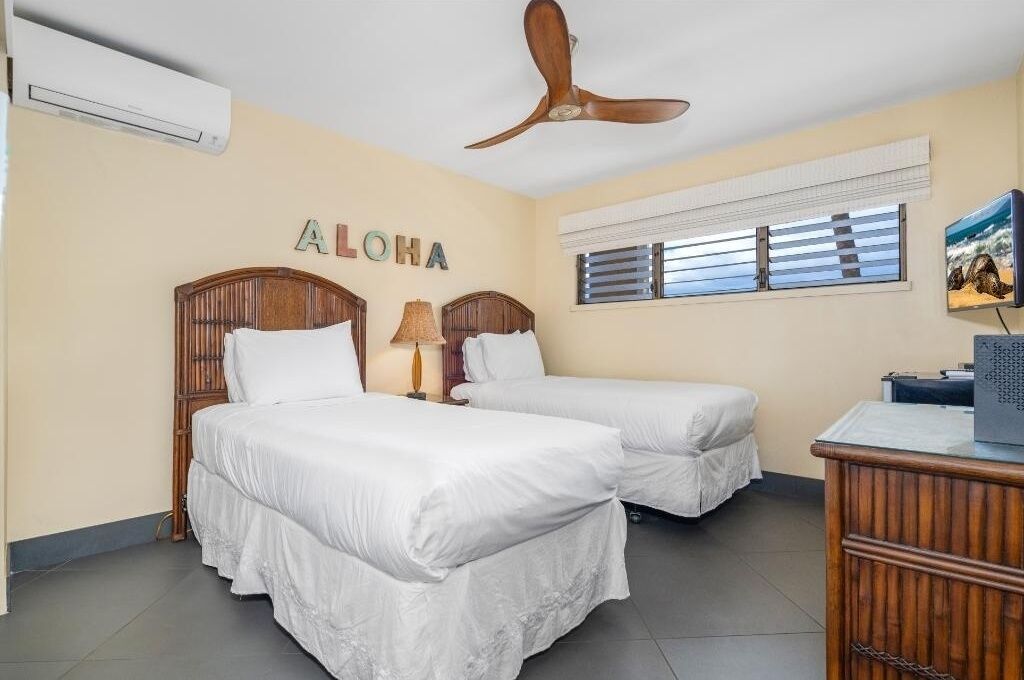 Een hotelkamer met twee bedden, een plafondventilator, een airconditioningapparaat, een raam met zonwering en een televisie op een houten dressoir.