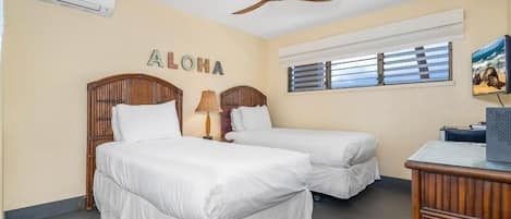 Een hotelkamer met twee bedden, een plafondventilator, een airconditioningapparaat, een raam met zonwering en een televisie op een houten dressoir.