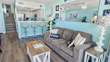 A living room with a grey sofa, a wooden coffee table, and a kitchen area with a bar stool set.