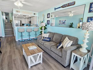 A living room with a grey sofa, a wooden coffee table, and a kitchen area with a bar stool set. - Oceanfront Second Floor Condo on the Boardwalk with Pool. Steps from the Ocean! El Capitan 204 (Ocean City)