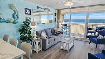 A living room with a view of the ocean, a ceiling fan, and a dining area with chairs and a table.