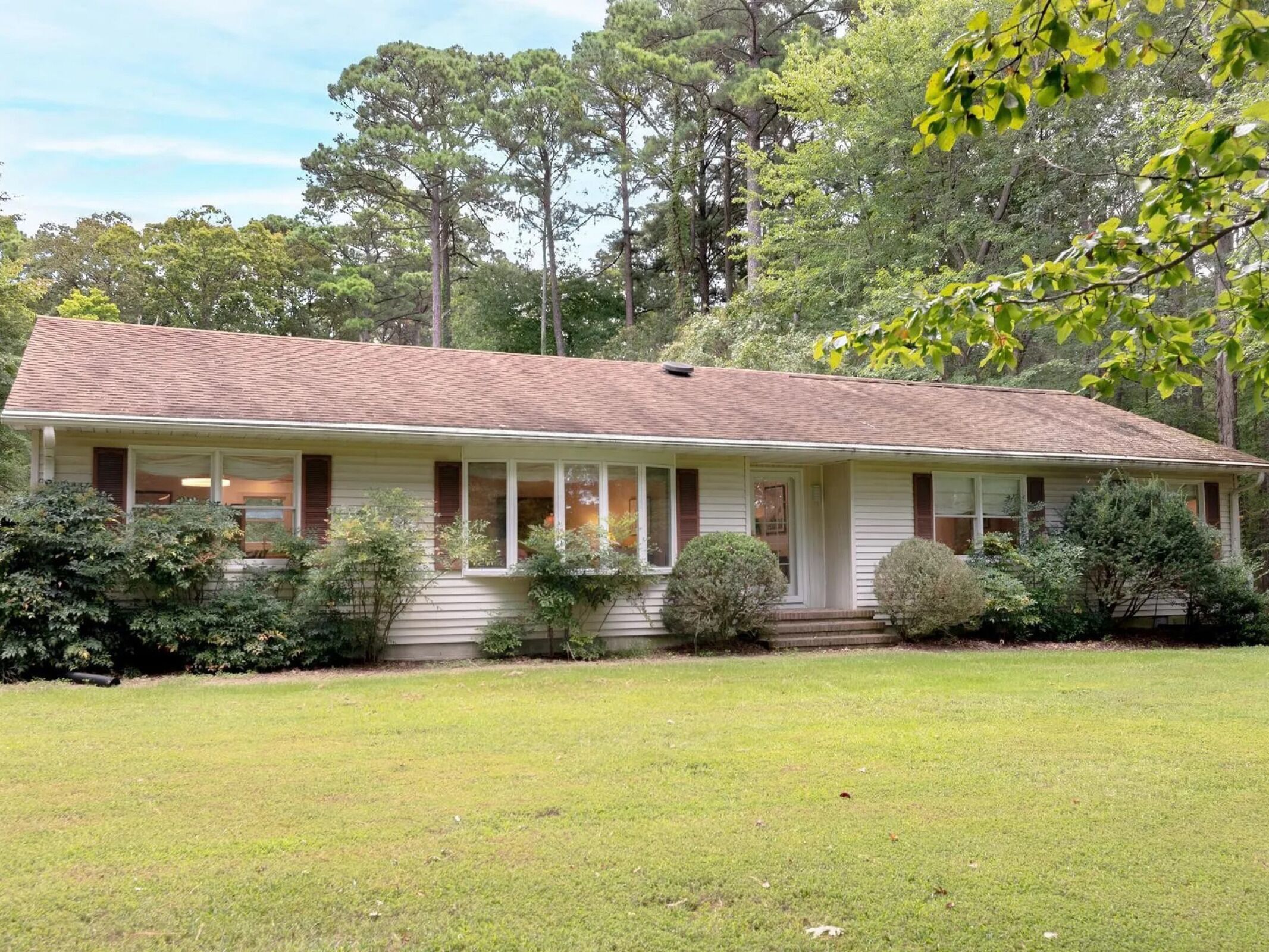 A charming ranch-style home surrounded by towering pines and lush landscaping, nestled on spacious grounds in a peaceful wooded setting.