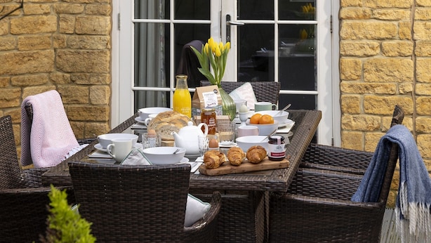 A breakfast setup with bread, pastries, and orange juice on a wicker table outside a stone building. - Appledore Cottage  - Comfy and cosy dog friendly holiday home in the Cotswolds (Andoversford)