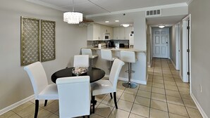 A dining area with a round table and white chairs, a kitchen in the background, and a hallway leading to a door. - Ocean Breeze West #504 | Gulf-Front Balcony + Resort Amenities (Perdido Key)