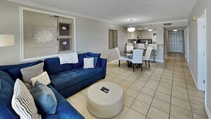 A living room with a blue sofa, a round ottoman, and a dining area with a table and chairs. - Ocean Breeze West #504 | Gulf-Front Balcony + Resort Amenities (Perdido Key)
