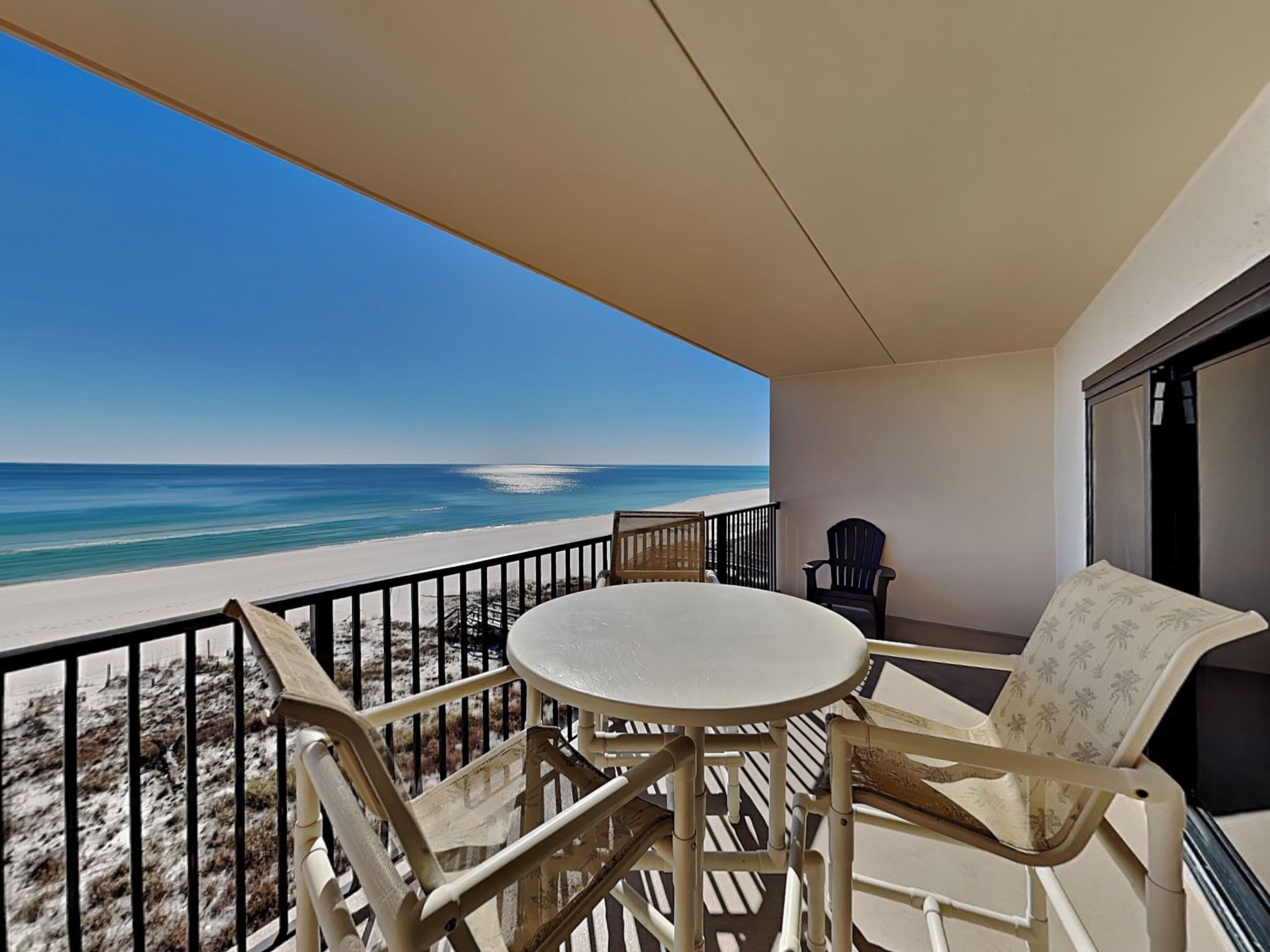 A balcony with a table and chairs, overlooking a beach with a clear view of the ocean.