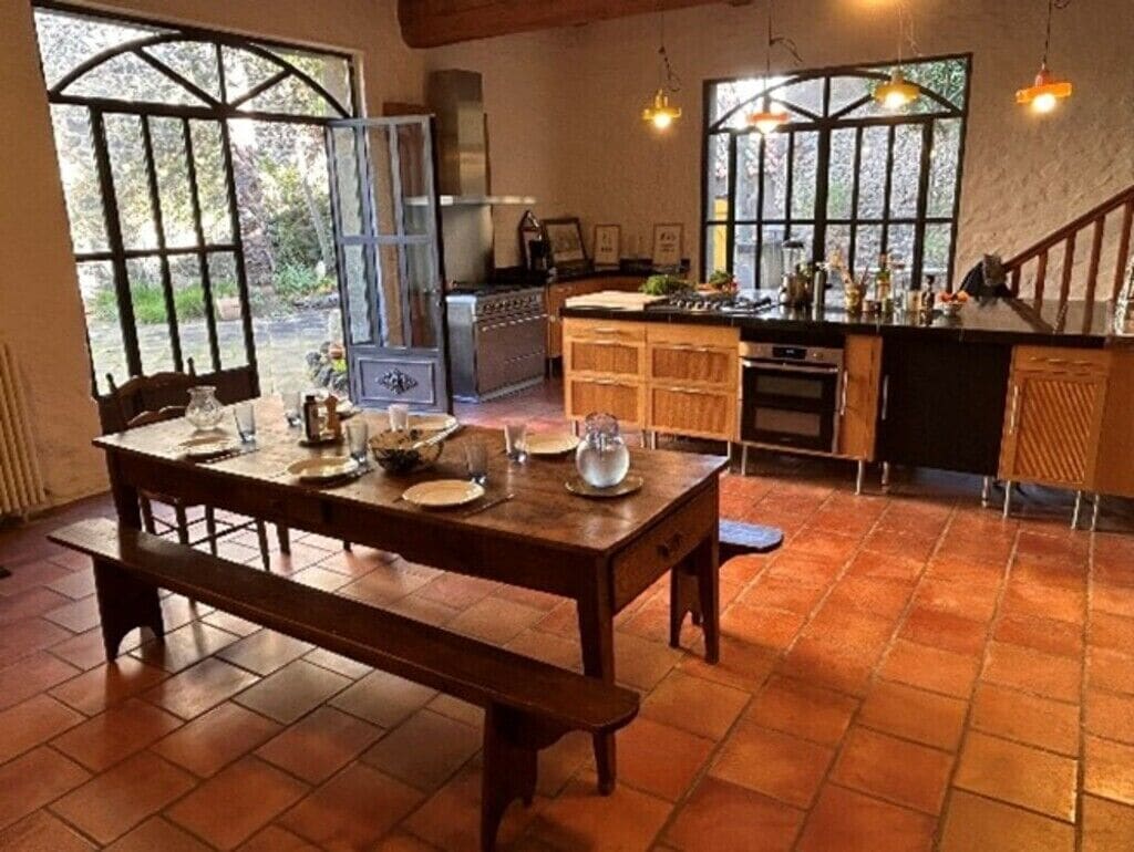A rustic kitchen with a wooden table set for dining, a stove, and a large window overlooking a garden.
