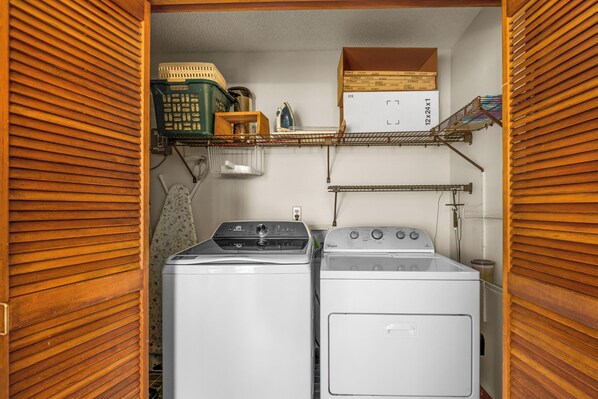 A laundry room with two stacked washers and dryers, wooden blinds, a shelf with boxes, and a small ironing board. - 🛥️🌴Canal-Front w/ Boat Dock | Two Primary Suites | Blue Heron Hideout (Pensacola Beach)