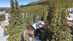 Eine Holzhütte, umgeben von hohen Nadelbäumen, mit freiem Blick auf eine kurvige Straße und ferne Berge.