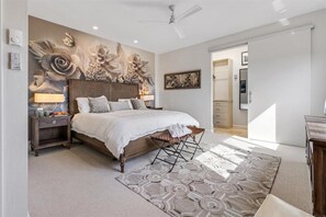 A bedroom with a large bed, a nightstand, a wall-mounted lamp, a decorative wall mural, a ceiling fan, and a door leading to another room.