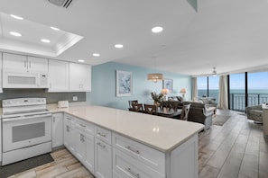 A modern kitchen with white cabinetry, a large island, and a dining area with a view of the ocean. - Condo with Ocean Views & Private Balcony, Near Pier (Garden City Beach)