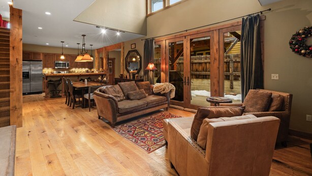 A spacious living area with a wooden floor, a sofa, a coffee table, and a dining table. There is a kitchen in the background with a refrigerator and a microwave. - Town Center Retreat | Hot Tub | Grill | 10 min to resort (Big Sky)