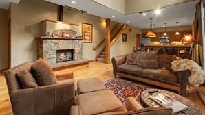 A spacious living room with a stone fireplace, leather sofas, and a wooden staircase leading to the kitchen. - Town Center Retreat | Hot Tub | Grill | 10 min to resort (Big Sky)