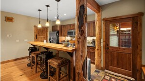 A rustic kitchen with wooden cabinets, a wooden bar counter, and hanging pendant lights. - Town Center Retreat | Hot Tub | Grill | 10 min to resort (Big Sky)