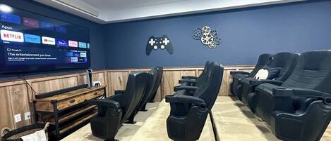 A cinema room with multiple black reclining seats, a wooden bench, and a large screen displaying streaming apps.