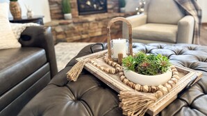 Una sala de estar acogedora con un sofá de cuero, una mesa de centro de madera con una vela y plantas, y una chimenea de piedra al fondo.
