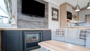 A modern living room with a fireplace, a flat-screen TV mounted on a wooden wall, a light-colored sofa, and a kitchen area with wooden cabinets and pendant lighting. - Seton Sands Gullane Luxe Lodge 3BR Sleeps 8 | WiFi (Port Seton)