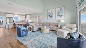 En rummelig opholdsstue med en stor sofa, et sofabord og en blå lænestol. I baggrunden er der et køkken med moderne hårde hvidevarer.