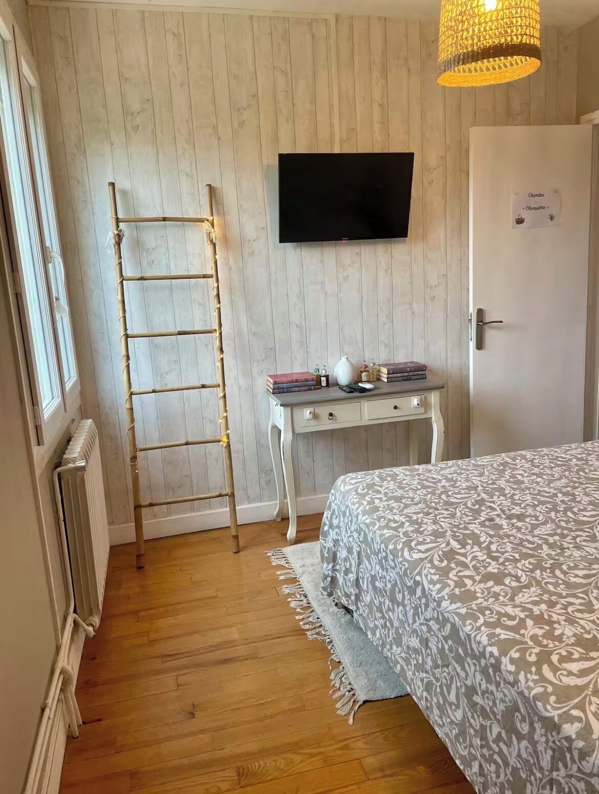 Une chambre avec un plancher en bois, un lit recouvert d’une couvre-lit à motifs, un bureau avec des livres et une lampe, un téléviseur fixé au mur et une échelle appuyée contre le mur.