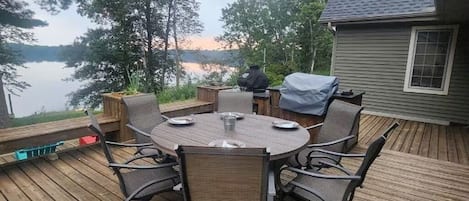 A wooden deck with a round table and chairs, overlooking a lake.