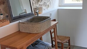 A bathroom with a stone wall, a large mirror, a vessel sink, wooden shelves, a chair, and a window with a view of trees.