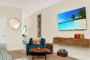 A living room with a teal sofa, a coffee table, a mounted TV displaying a beach scene, and a wooden shelf with books and decorative items.