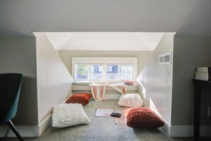 Un pequeño dormitorio en el ático con techo inclinado, una ventana y vistas al exterior. Hay almohadas y un juego en el suelo.
