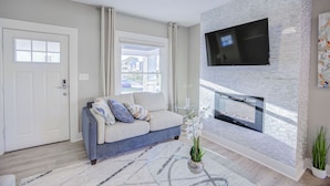 A modern living room with a sofa, a fireplace, a flat-screen TV mounted on a textured wall, and a ceiling fan. - Cozy Fountain Square Retreat Just Steps from Downtown Indianapolis (Indianapolis)