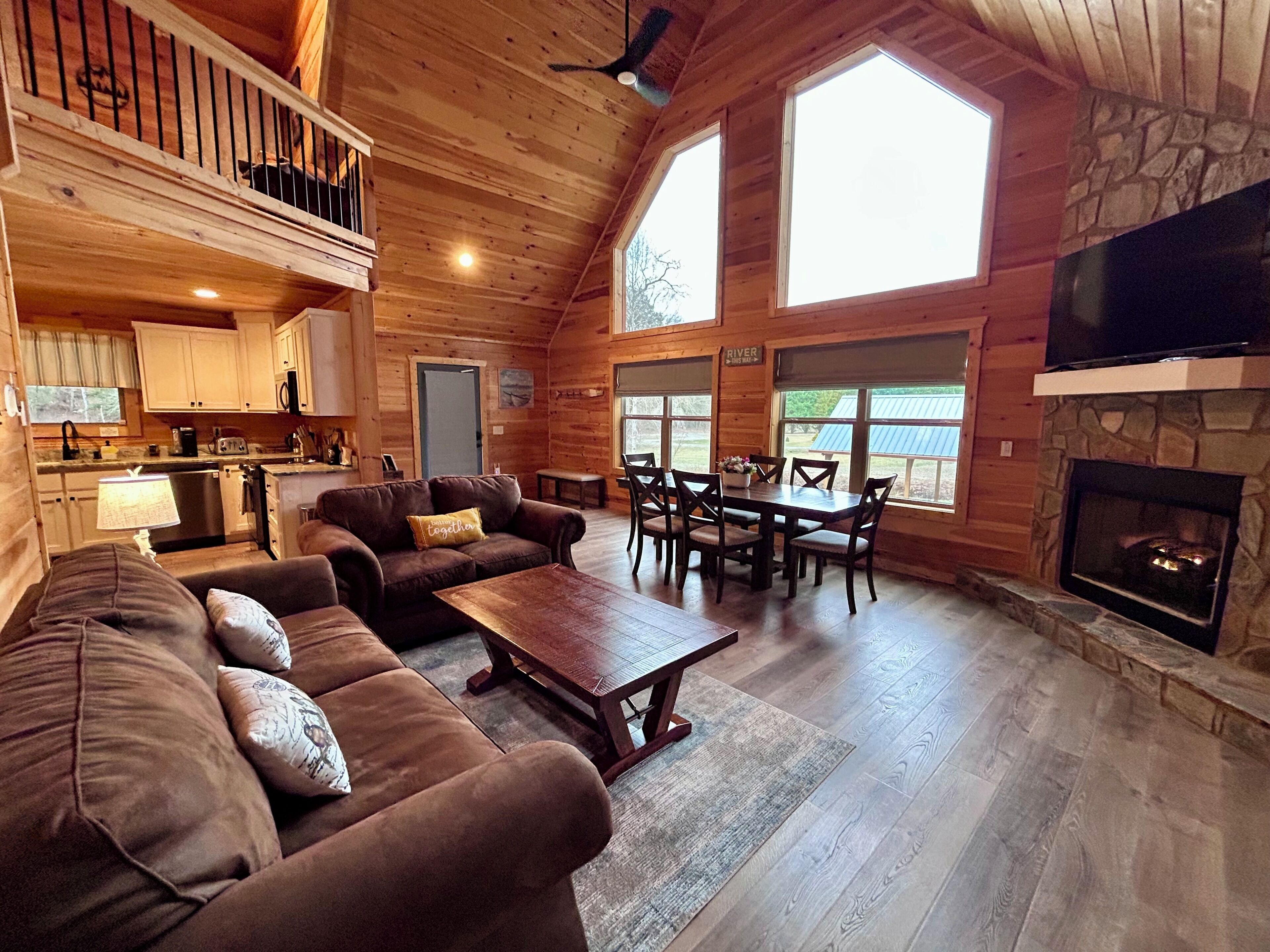 A cozy cabin interior with a living room, dining area, kitchen, and a stone fireplace.