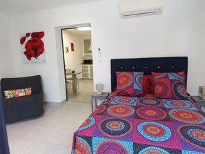 A bedroom with a bed, a sofa, and a kitchen visible through an open door. - GITE D'EMMA EN PROVENCE 2 PEOPLE AND 1 CHILD (Camaret-sur-Aigues)