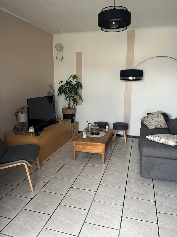 A living room with a TV on a wooden cabinet, a coffee table with decorative items, a sofa, a potted plant, and a hanging light fixture. - Cozy Apartment   (Pertuis)