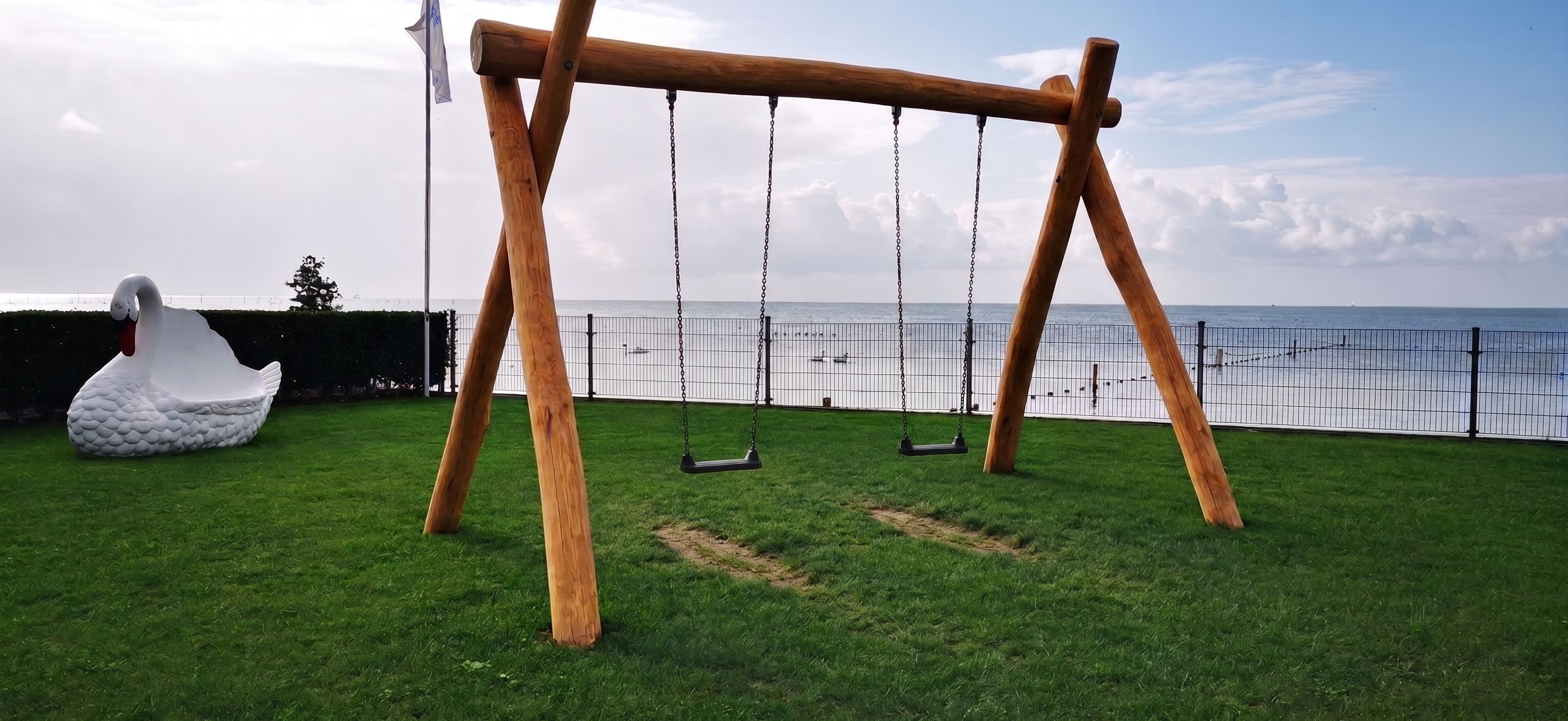 Un ensemble de balançoires en bois, agrémenté d’une sculpture de cygne, installé sur une pelouse verdoyante et offrant une vue sur une étendue d’eau.