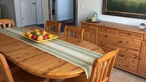 Une table à manger en bois, recouverte d’une nappe à rayures, est entourée de chaises en bois. Un panier de fruits est posé sur la table.