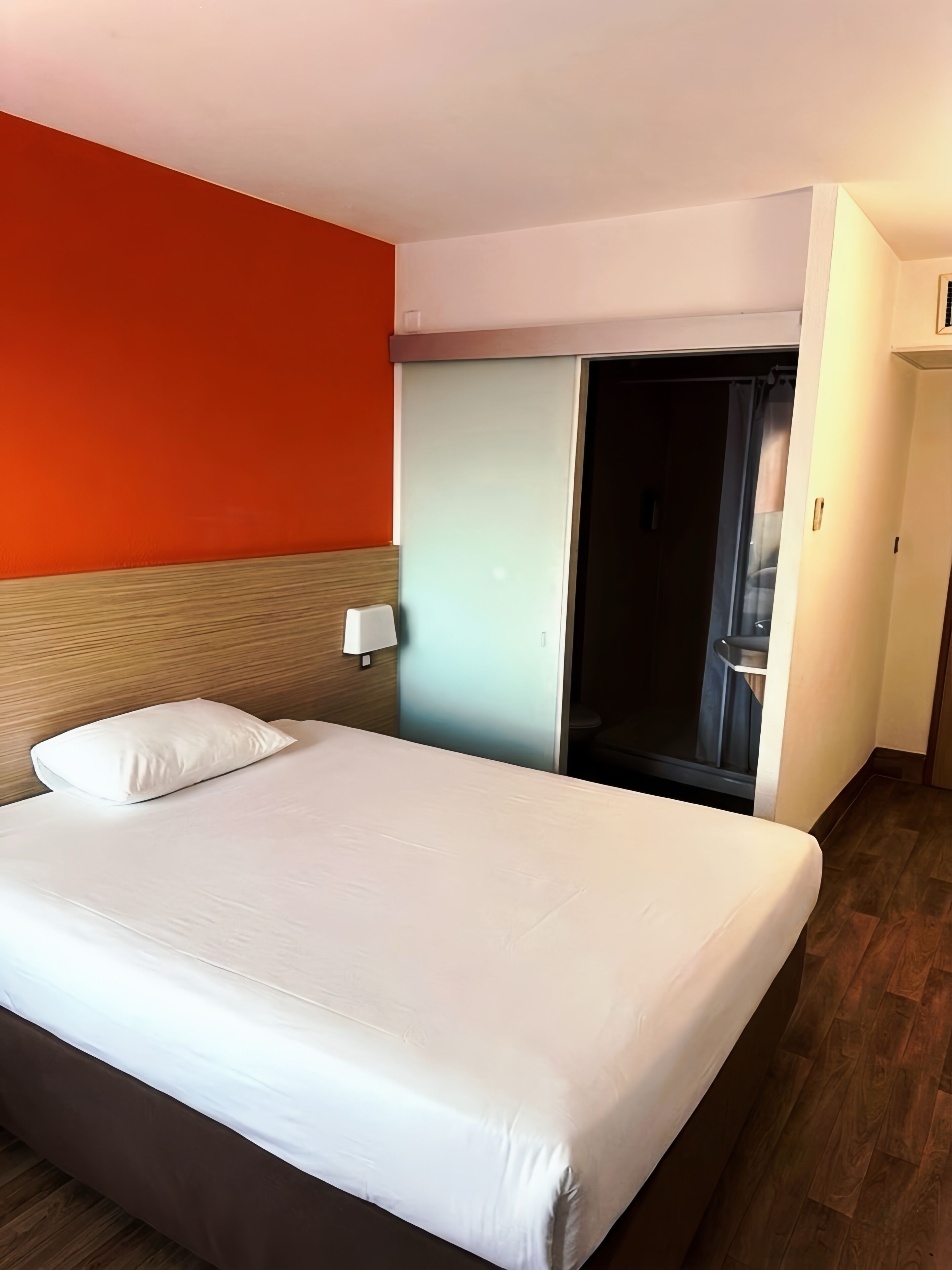 A hotel room with a bed, a nightstand, a wall-mounted lamp, and a sliding door leading to a bathroom.