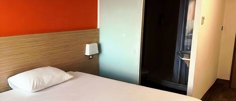 A hotel room with a bed, a nightstand, a wall-mounted lamp, and a sliding door leading to a bathroom.