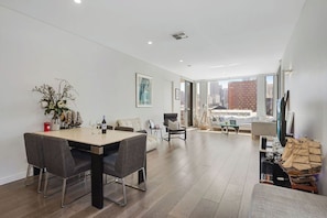 A spacious living area with a dining table, chairs, a sofa, and a large window offering a city view. - Lux 2BR apartment in Darling Harbour with Balcony Spa (Ultimo)