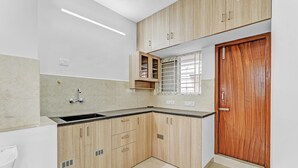 A kitchen with wooden cabinets, a sink, and a window with blinds. - Ecostay Valasaravakkam (Chennai)