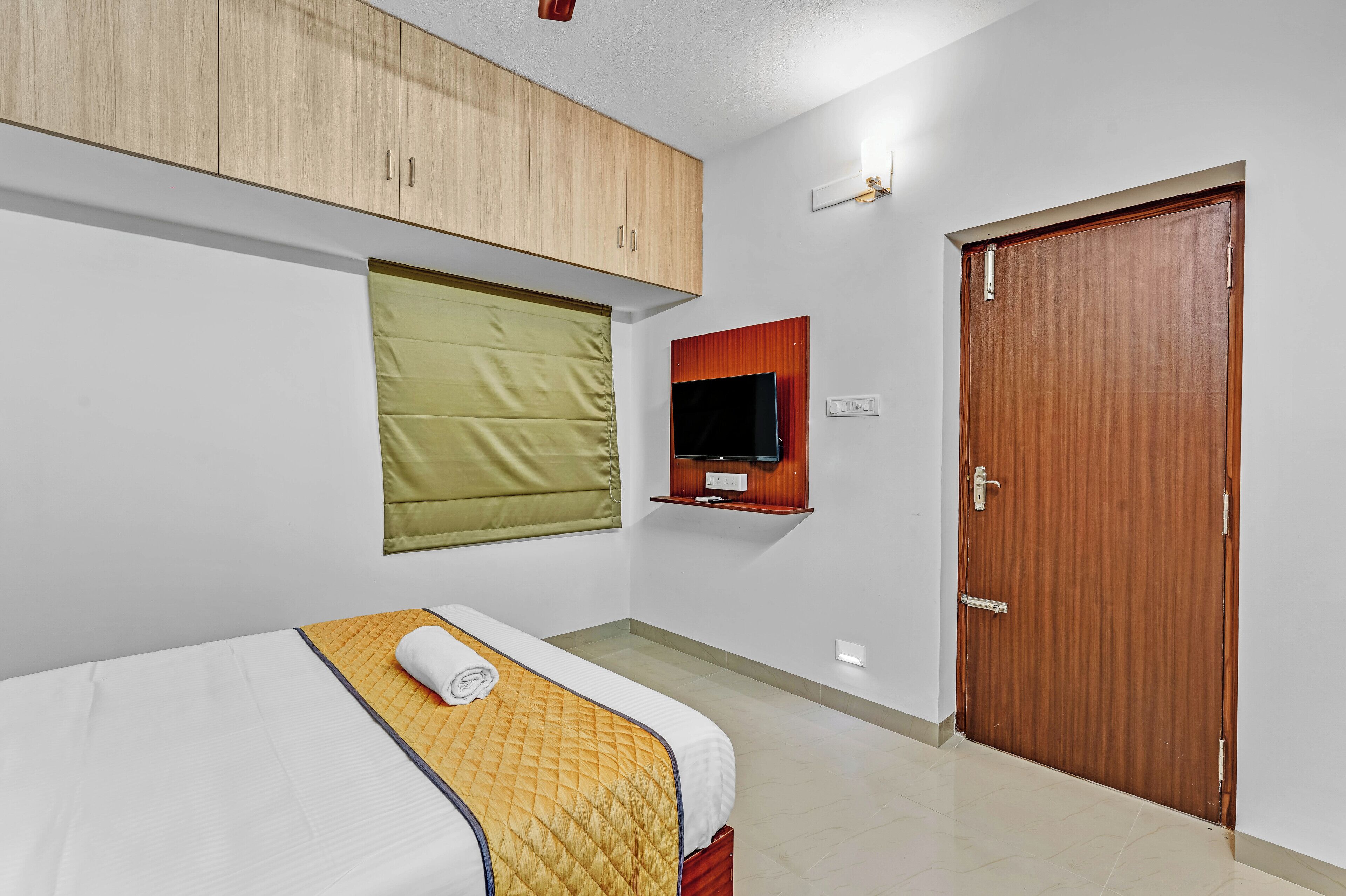 A hotel room with a bed, a television mounted on the wall, a wooden wardrobe, and a door.