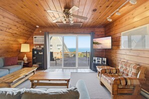 Ein Interieur einer Holzhütte mit Holzbalkendecke, Deckenventilator, Fernseher, einer Couch, einer Bank und Blick auf den Ozean durch eine Glastür.