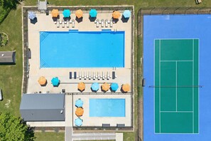 Eine Vogelperspektive auf einen Poolbereich mit Tennisplatz, mehreren Sonnenschirmen und Liegestühlen.